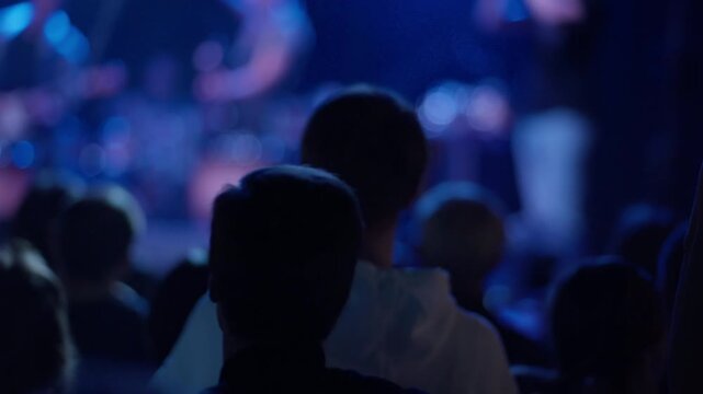 Fan raises clasped hands at concert while another arm extends beside, blurred guitarist with guitar visible on stage in blue light