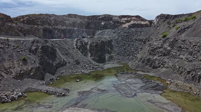 A large mining site displays deep excavated pits filled with water. The landscape shows gray rock formations and a cloudy sky in the background. Machinery is visible in the distance.