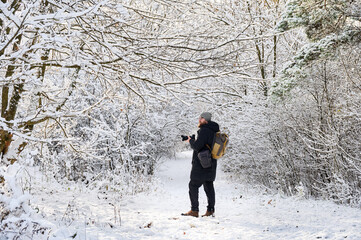 Fototapeta premium Winter photographer stands on snowy forest path with camera, surrounded by frosted branches and quiet woodland. Useful for travel marketing, winter tourism, outdoor advertising, and lifestyle concepts