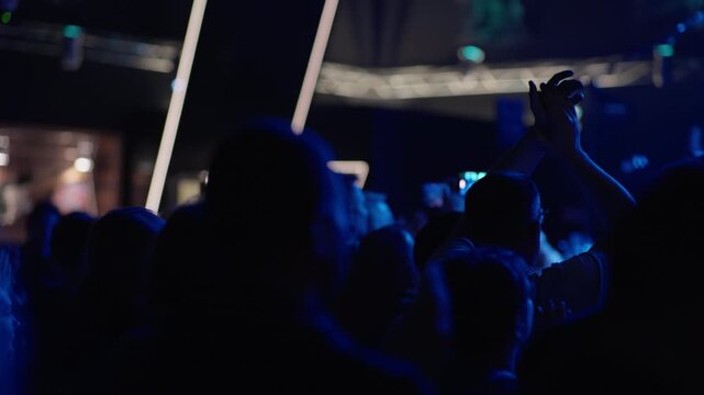 Fan holds smartphone raised to film live performance, performer dimly visible on stage at back, blue atmosphere, surrounding crowd in dark