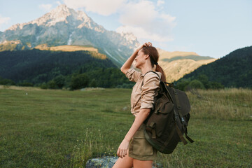 Naklejka premium Young woman with backpack enjoys sunlit meadow as distant mountains rise, capturing a candid moment of adventure, freedom, and connection with nature.