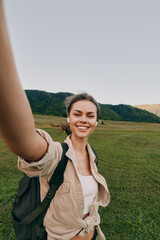 Naklejka premium Cheerful woman outdoors, smiling while taking a selfie in a sunlit field, casual jacket, backpack, relaxed pose, natural colors, expansive landscape behind and light adds warmth