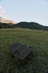 Naklejka premium A lone backpack lies on a grassy meadow with distant hills and a clear sky, conveying outdoor travel, exploration, and a sense of nature escape.