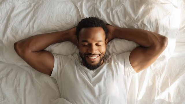Top view of a happy African American man waking up and stretching in bed with white sheets in morning sunlight.
