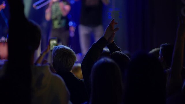Audience enjoying live music concert. People clap hands in dark venue with blue stage lights. Band performs in background. Shot from behind crowd shows energetic atmosphere at festival
