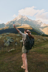 Naklejka premium A woman with backpack hikes through alpine meadow, arms raised, rugged mountains in the background under a bright sky, conveying adventure and outdoor lifestyle.