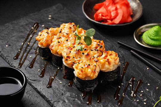 Baked warm sushi roll topped with sesame seeds and micro greens on black slate plate with soy sauce, wasabi and ginger over black concrete background. Close up view 
