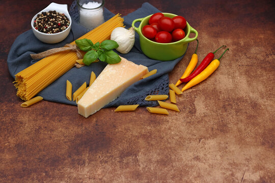 Pasta ingredients: Spaghetti, penne, Parmesan, garlic, basil, colorful chili peppers, cherry tomatoes in a green pot, pepper and salt shakers on a brown stone base.