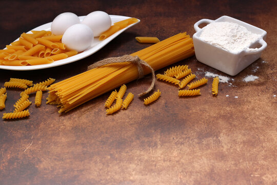  Ingredients for homemade pasta: Various uncooked pasta shapes with eggs, flour and noodles on a stone-look background with space for text.
