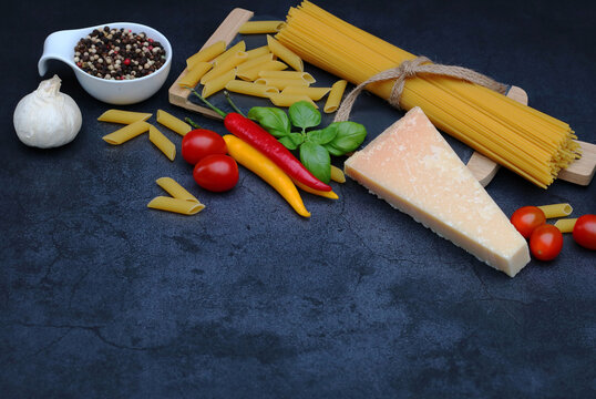 Italian pasta ingredients including spaghetti, Parmesan, chili and herbs on a dark stone background with space for text.