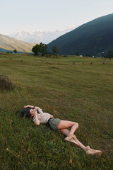 Naklejka premium Relaxed woman lying in a green meadow with distant hills, enjoying nature and sunshine. Casual outfit highlights outdoor lifestyle and a peaceful, carefree moment and mood.
