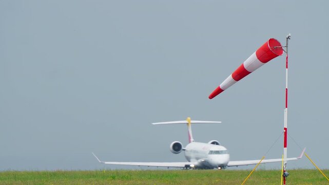 Red and white striped windsock extended in strong wind on pole at airport with passenger aircraft taking off in background on taxiway, overcast sky