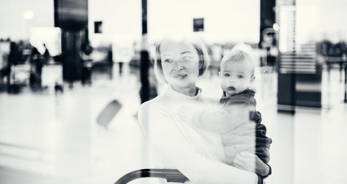 Thoughtful young mother looking trough window holding his infant baby boy child while waiting to board an airplane at airport terminal departure gates. Travel with baby concept