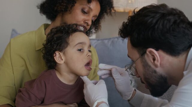 Zoom out shot of young multiethnic doctor in white coat using tongue depressor while examining sore throat of small boy sitting with African American mother during home appointment