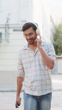 Happy overjoyed young man tourist in wireless headphones choosing, listening favorite energetic disco music on smartphone outdoors. Indian tourist guy walking passes by urban sunny city town street.