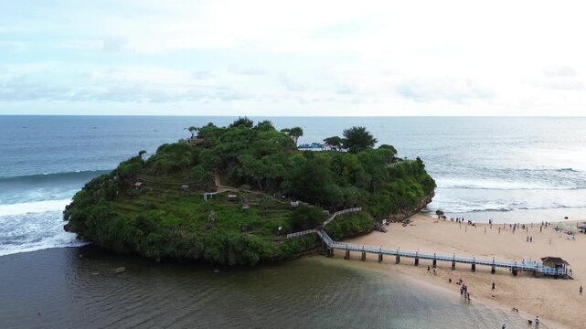 Aerial drone view of Drini Beach in Yogyakarta, Indonesia. Scenic tropical coastline with white sandy beach, clear turquoise water, small offshore island, and gentle ocean waves.