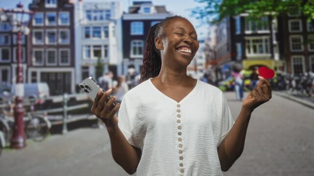 Young african american woman wearing white blouse, holding smartphone and small red paper heart with raised hands on a busy street, smiling and laughing; joy connection.