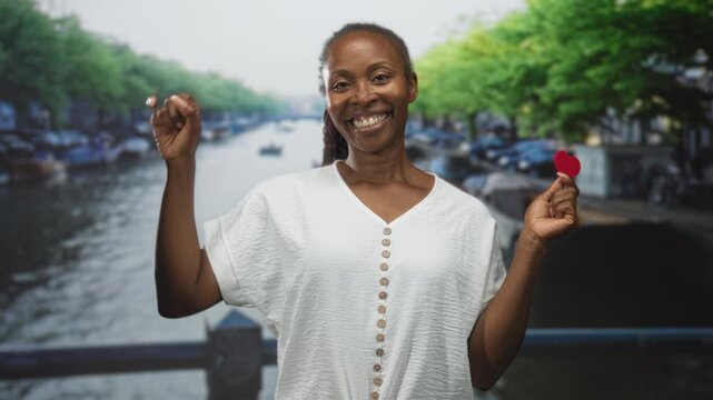Woman smiling broadly holds small red heart between thumb and forefinger while standing on a street bridge over a canal, wearing a white button blouse; love joy connection.