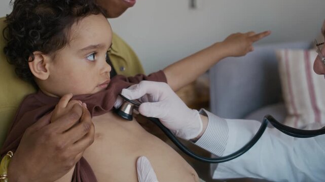 Medium close up handheld shot of male medical worker in gloves and white coat listening to breath of African American toddler with stethoscope, sitting with mother during home visit