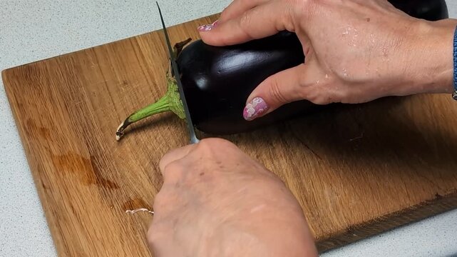 Purple eggplant placed on a wooden cutting board next to a sharp kitchen knife. A hand with manicured nails gently touches the eggplant, emphasizing food preparation and fresh ingredients commonly.