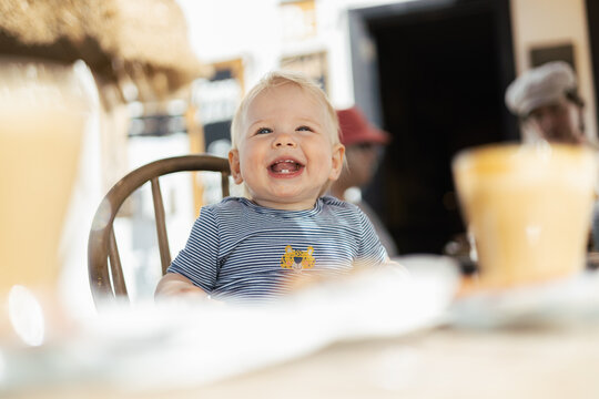 Adorably happy blonde toddler smiling brightly at the camera in his high chair. Joyful child laughing
