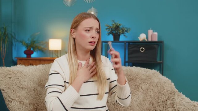 Caucasian young adult woman sitting on cozy sofa at home, holding smartphone, looking shocked and disappointed after reading bad news or losing lottery. Emotional girl indoors with modern decor
