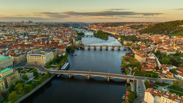 Aerial view of Charles Bridge crossing the Vltava River surrounded by historic red rooftops and lush green hills under a golden sunset in Prague, Prague, Czechia.