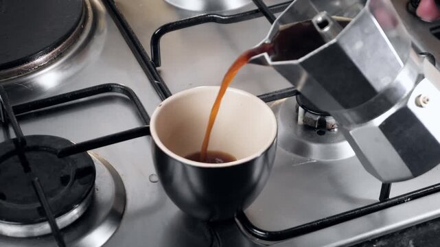 Pouring fresh coffee from a moka pot into a black cup on a gas stove