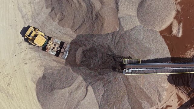 Heavy machinery is operating at a sand and gravel site. A scraper is moving material while a conveyor is transporting it. Dust is rising from the ground. The sun is shining.
