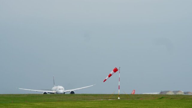 Windsock leaning horizontally in strong wind on airfield with wide body aircraft taking off in background, green grass and gray overcast sky