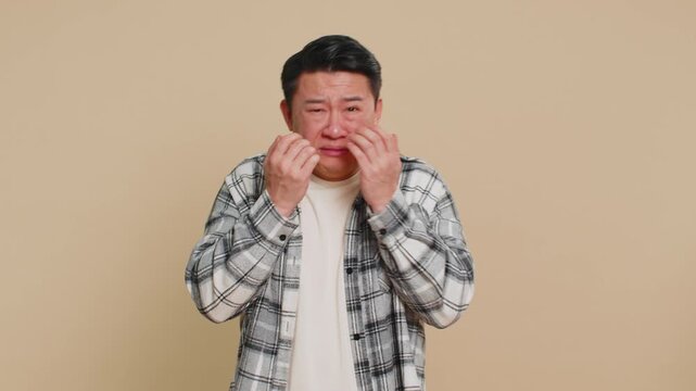 Upset disappointed Asian middle-aged man wipes tears and cries from despair, being sad because of unfair problem things, fail, lose, bankruptcy. Mature Chinese guy isolated on beige background indoors