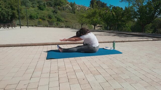 A woman performs yoga and stretching exercises outdoors on a mat in a park. This scene captures her dedication to fitness, flexibility, and overall wellness in the natural environment.