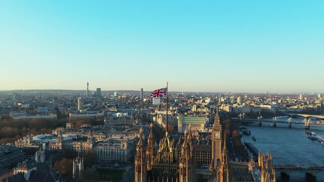 Waving Union Jack flag on top of Victoria Tower in London with Big Ben and River Thames while camera orbits the Houses of Parliament on a winter sunset