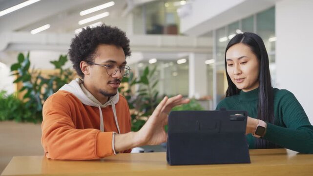 Young coworkers couple discussing something about what they see on tablet screen. Male smiling. Asian woman explaining details with gestures. Completing task together in office.