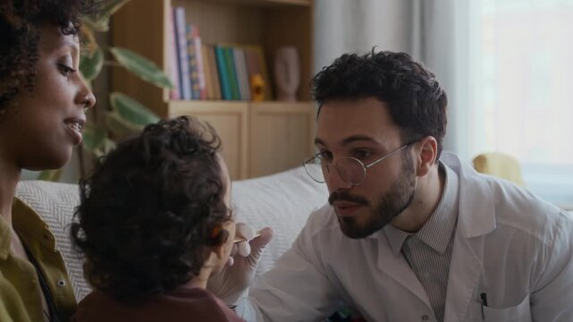 Chest up shot of male paediatrician examining throat of small patient with tongue depressor during appointment at home