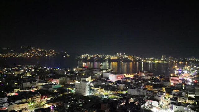 Captivating aerial drone video of Acapulco Bay, Mexico, at night. The vibrant city lights illuminate the coastline and reflect beautifully on the dark ocean waters, showcasing an impressive urban pano