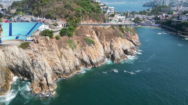 Lateral drone video capturing the picturesque coastline of Acapulco, Mexico, with its rugged cliffs, sparkling blue ocean, and distant city.
