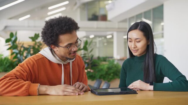 Excited colleagues giving high five after successfully completing task at work. Young African American man in orange hoodie smiling to his Asian friend. Tablet laying on the table.