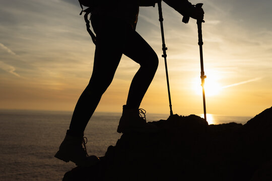 Hiker in silhouette trekking up a rocky ocean cliff at sunset, symbolizing challenge, determination, success, and overcoming obstacles to reach goals
