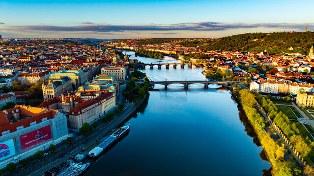 Aerial view of the Vltava River and historic bridges including Charles Bridge under a golden sunset light with red-roofed buildings and lush greenery in Prague, Prague, Czechia.