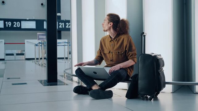 Young male passenger with curly hair sitting on the airport floor using a laptop while waiting