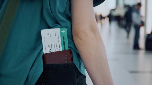 An unrecognizable female traveler walks through a modern airport terminal building, with a close-up showing her passport and boarding pass tucked into the back pocket of her jeans.