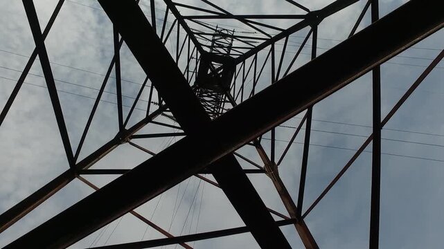 Low-angle view looking up through a steel lattice high-voltage transmission tower with power lines and insulators against a cloudy sky