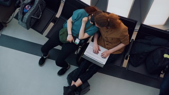 A top-down view shows a tired woman resting peacefully in an eye mask on her partner's shoulder, as he works efficiently on his computer. 