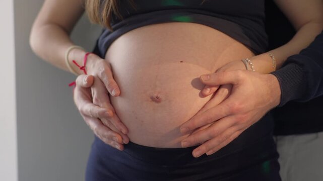 Pregnant woman belly held gently by two sets of hands from both sides, close frame showing skin texture and gentle touch, green bokeh visible