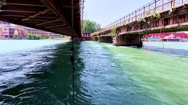 The teal waters of the Ganges flow rapidly between two parallel iron bridges in Haridwar, with an iron chain dangling from the left structure and sunlit buildings lining the distant riverbank.