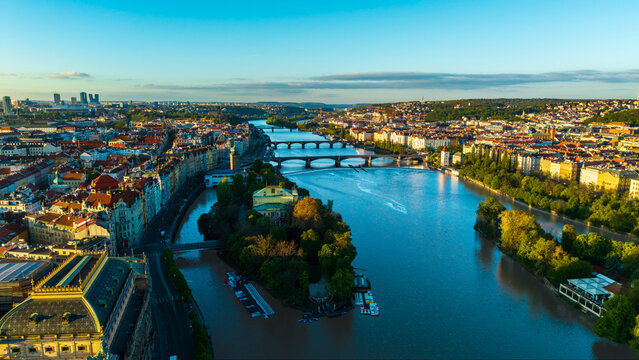 Aerial view of the Vltava River flowing through the historic city center with multiple bridges and red-roofed buildings under a clear blue sky in Prague, Prague, Czechia.