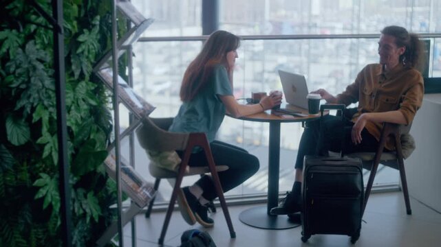 A young couple sits at a cafe table in a bright airport terminal, surrounded by travel essentials: laptop, passports, boarding passes, and coffee. 