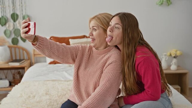 Woman and daughter hold smartphone taking selfie on bed in building, mother makes duck face while daughter sticks out tongue; playful bonding.