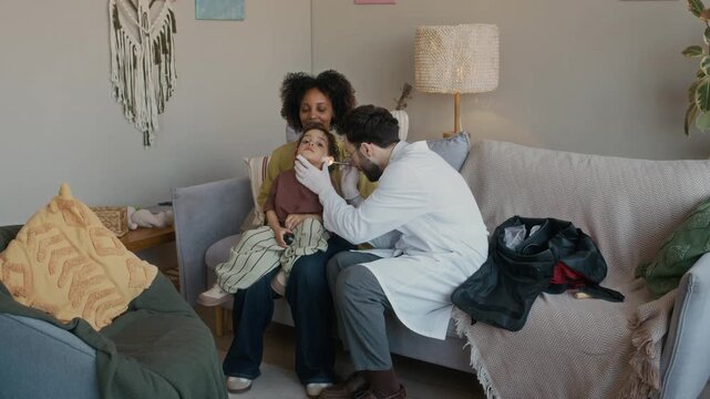 Long shot of multiethnic pediatrician using flashlight while examining ears of small patient sitting on mothers lap during appointment at home